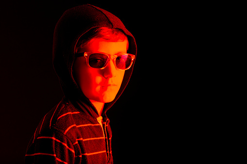 Caucasian child with hooded sweatshirt and sunglasses, with serious expression, illuminated with a red light, on black background and space to copy.