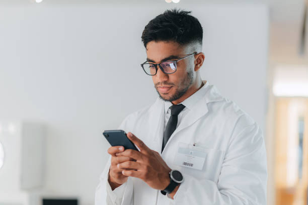 Doctor, phone and break with medical healthcare expert reading message, social media notification and online news. Man medical worker or gp using a mobile app with 5g wifi network connection at work stock photo
