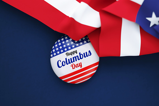 Happy Columbus Day written badge sitting behind rippled American flag over dark blue background. Horizontal composition with copy space. Directly above.