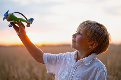 Happy child playing with a toy plane in nature during summer sunset. Boy in a  white shirt with a plane in hands on wheat field. Kid holds a wooden airplane and dreams of being a pilot, on the nature