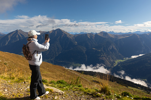 An adult woman in a sports jacket, trousers and a cap photographs a landscape in the Austrian Alps. High quality photo