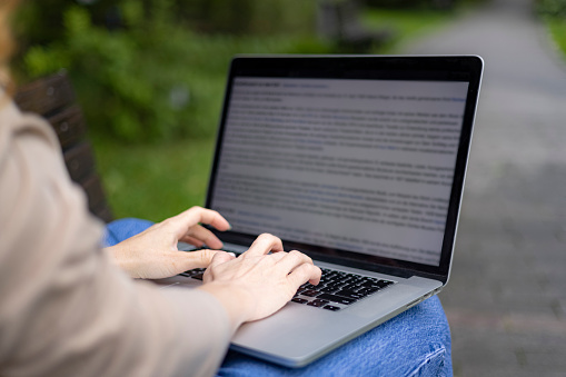 Close up of an unrecognizable woman sitting outdoors, typing on a laptop