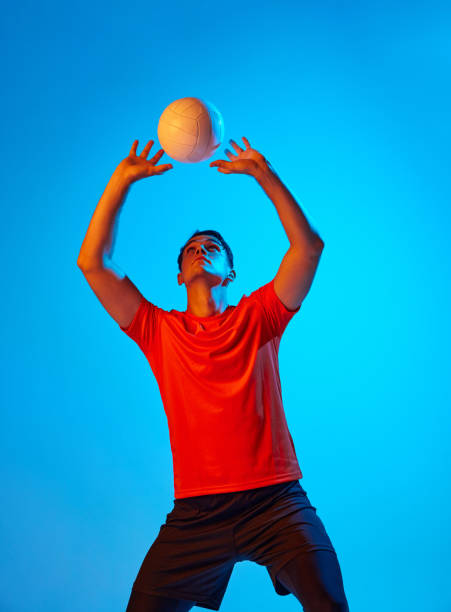 One male volleyball player in orange-black uniform training with ball isolated on blue studio background in neon light. Sport, gym, team sport, challenges