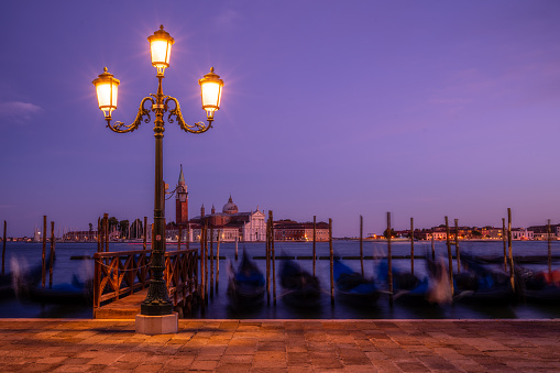 Old street lamp shining on empty waterfront with gondolas parking in Grand Canal in Venice, facing opposite side with Campanile di San Giorgio on quiet evening, long exposure shot