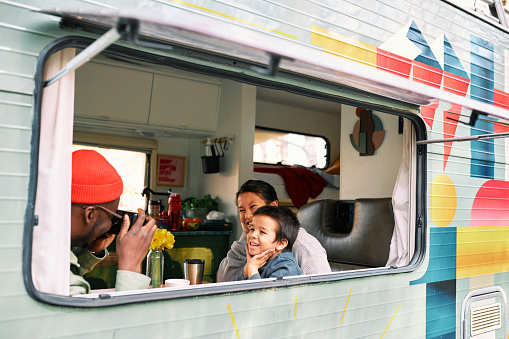 An African American father photographs his son and Asian wife in a van. They are sitting around the table in the comfy van they live in.