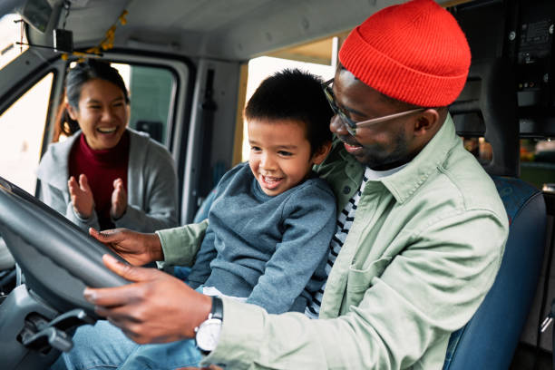 A nomadic family travels in their motorhome. A multicultural father holds his toddler son in a lap and drives their home on the wheels while a mother applauds and looks at them with a smile on her face. family-van-driving stock pictures, royalty-free photos & images