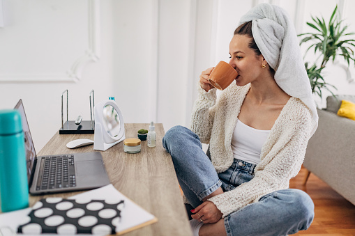 One woman, beautiful young woman at home, watching a movie on laptop and holding a cup of coffee, she has a towel on her head.