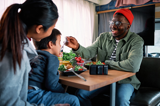 A cheerful African father feeds his son across the table in their van with healthy food. The Asian mother holds her son and smiles.