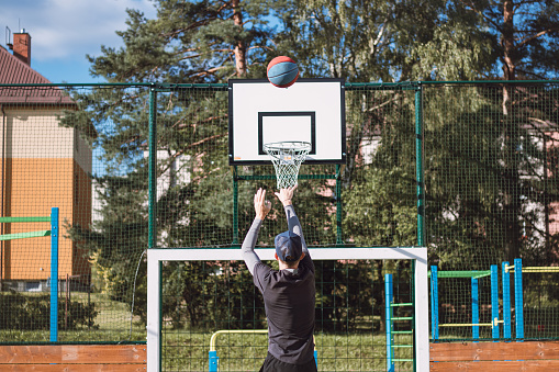 Blond boy in sportswear practices shooting a basketball from behind the three-point line. Outdoor basketball court. Preparing for the season.