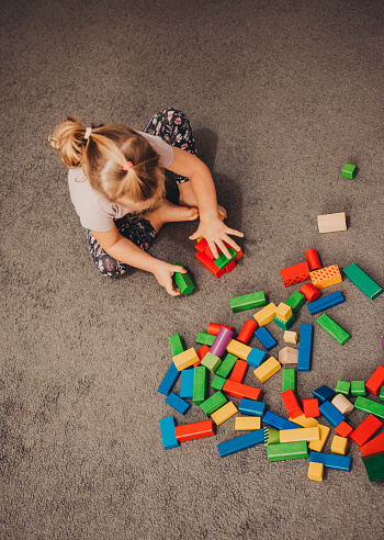 Cute baby girl is building tower with toy blocks on the ground. Indoor.