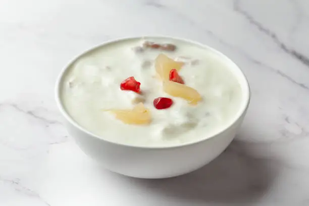 Close-up of Fruits raita or Yogurt & Pineapple or Ananas Raita garnished with pineapple piece and pomegranate, served in a white ceramic bowl over whiet granite background