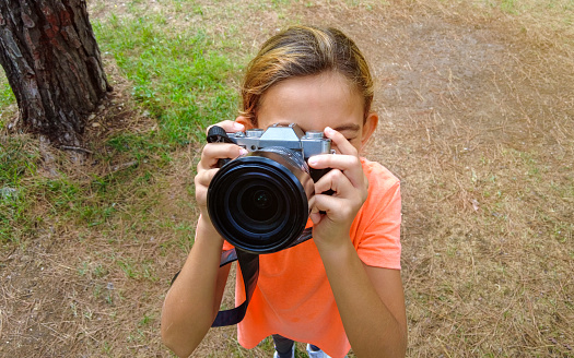 11-year-old girl holding a mirrorless camera. He's looking through the viewfinder trying to take a picture. From the waist up, front facing the camera. Shot from above. Background grassy field.