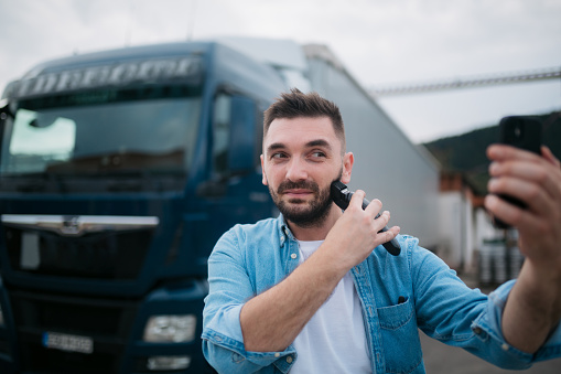 Male truck driver taking a break from driving, shaving his beard using an electric trimmer and a smart phone instead of a mirror
