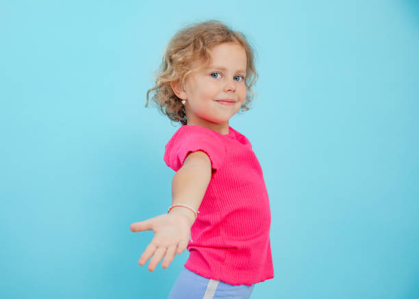 Nice little girl invite people and lend outstretched palm on empty blue background, copy space. Portrait of happy child of kindergarten age with pretty curls in casual wear. Invitation, follow me.
