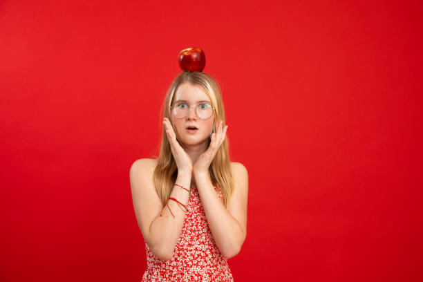 Open eyed teen girl stand with apple lying on head, empty red background, copy space. Portrait of clever child in glasses and floral print dress with surprise emotion. Idea, shock, science symbol Open eyed teen girl stand with apple lying on head, empty red background, copy space. Portrait of clever child in glasses and floral print dress with surprise emotion. Idea, shock, science symbol. gawp stock pictures, royalty-free photos & images