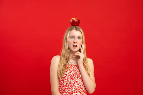 Open eyed teen girl stand with apple lying on head, empty red background, free copy space. Portrait of clever child in floral print dress with surprise emotion. Idea, study, shock, science symbol Open eyed teen girl stand with apple lying on head, empty red background, free copy space. Portrait of clever child in floral print dress with surprise emotion. Idea, study, shock, science symbol. gawp stock pictures, royalty-free photos & images