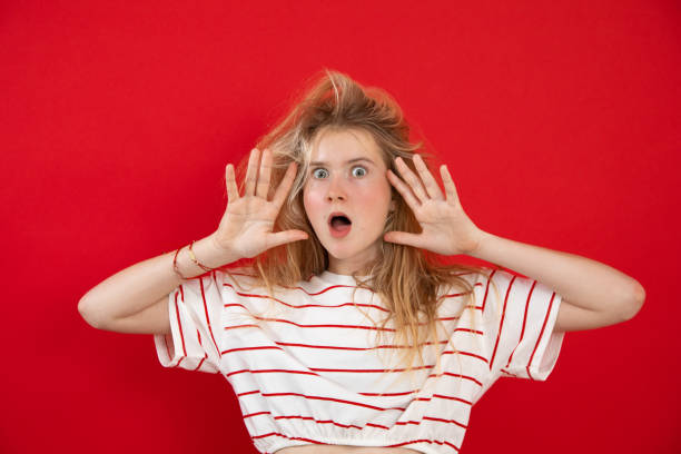 Open eyed young lady stand with raised hands with spread fingers near face on empty red background. Nice girl with wild hair have fun and show surprise emotion. Astonishment, wow effect Open eyed young lady stand with raised hands with spread fingers near face on empty red background. Nice girl with wild hair have fun and show surprise emotion. Astonishment, wow effect. gawp stock pictures, royalty-free photos & images