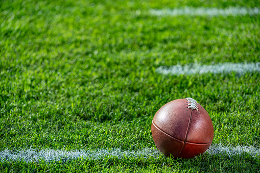 A low angle close-up view of a leather American Football sitting in the grass on a white yard line with hash marks in the background.