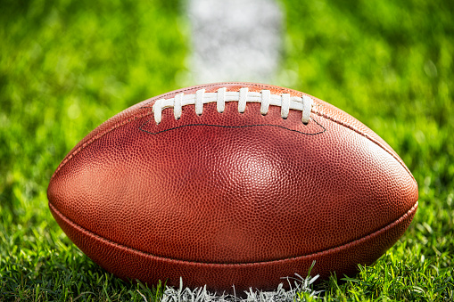 A low angle close-up view of a leather American Football sitting in the grass on a white yard line.