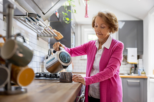 Senior woman at home making a cup of tea and pouring water from the kettle - domestic life concepts