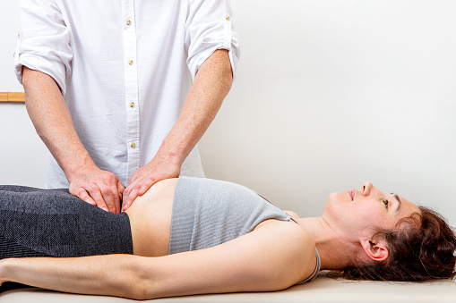 Male osteopath (chiropractor) healing a young woman lying down on a massage bed by touching her abdomen. Can illustrate the concept of Osteopathy, Alternative medicine, Physiotherapy, pain relief. Space for copy.