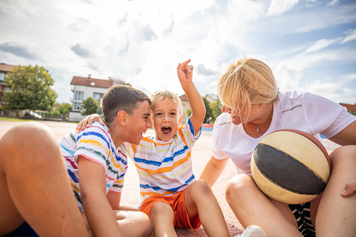 Portrait of Caucasian mother with her two son, sitting on the sports court and enjoy the active and playful summer day together