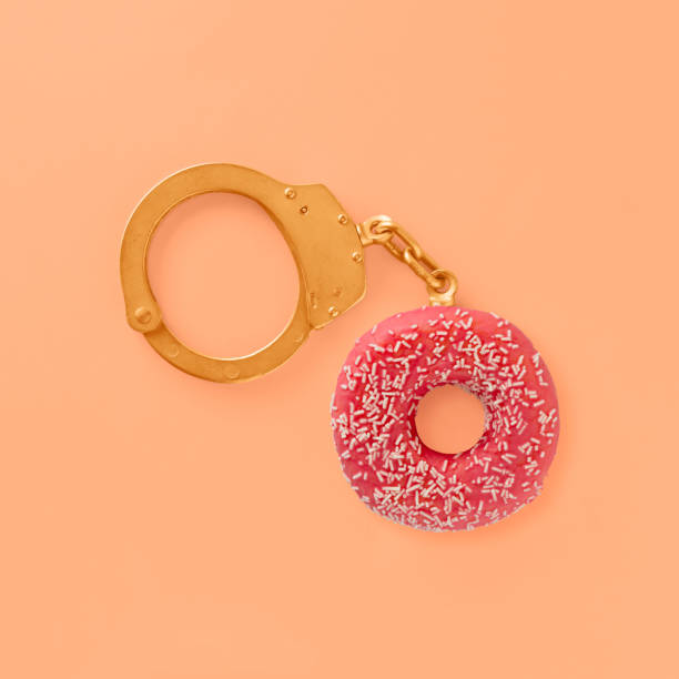 rosquilla rosa coral con destellos blancos como parte de esposas doradas sobre fondo naranja pastel aislado. adicción al azúcar o a los dulces. mínima estética abstracta plana tendida. concepto de hábitos alimenticios poco saludables. - golden handcuffs fotografías e imágenes de stock