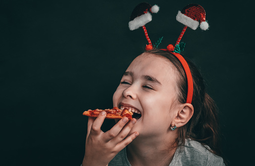 Happy little caucasian girl with closed eyes and a rim on her head made of santa claus hats holds in her hand a slice of pizza near her mouth on the right on a black background with copy space on the left, close-up side view.