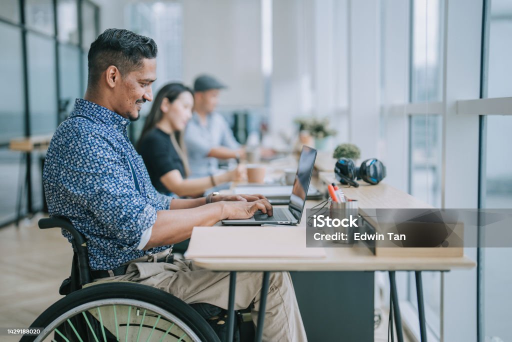 Asian Indian white collar male worker in wheelchair concentrating working in office beside his colleague Accessibility for Persons with Disabilities Stock Photo