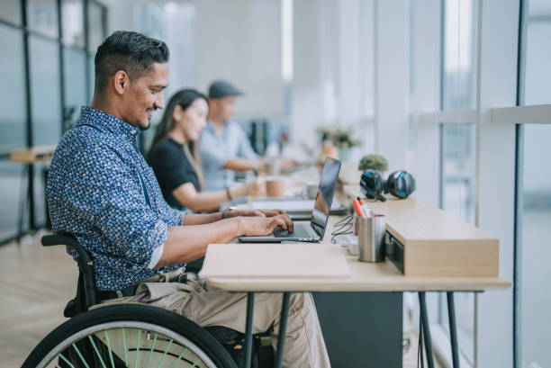 Asian Indian white collar male worker in wheelchair concentrating working in office beside his colleague stock photo