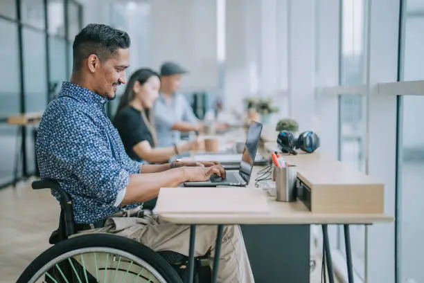 Photo of Asian Indian white collar male worker in wheelchair concentrating working in office beside his colleague