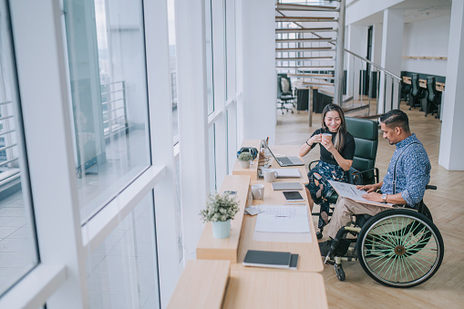 Indian white collar male worker in wheelchair  discussion  with  female Asian Chinese colleague coworking in creative office workstation beside window