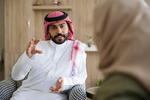 Saudi couple enjoying conversation while relaxing at home Personal perspective over the shoulder of Middle Eastern woman in traditional attire sitting face to face and talking with bearded man in early 30s. kaffiyeh stock pictures, royalty-free photos & images