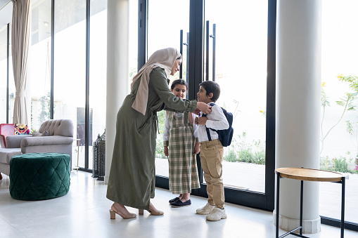 Full length view of late 20s woman in abaya and hijab standing at front door helping son with his backpack before he and his sister leave in the morning.