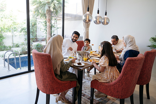 Wide angle view of men and women in traditional attire and children in western dress sitting around dining table in modern Riyadh home eating and talking.