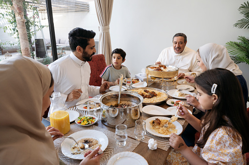 Middle Eastern men and women in traditional and western attire sitting around dining table in family home eating kabsa, harees, and saleeg.