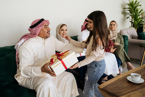 Mature Middle Eastern man in traditional attire interacting with his 6 year old granddaughter as the family enjoys social gathering and generosity.