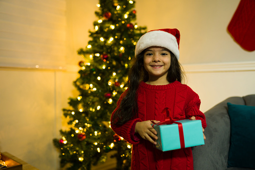 Adorable hispanic young girl wearing a santa hat at home holding a present in front of the christmas tree