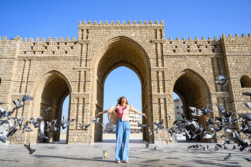 Full length front view of smiling young woman standing in front of historical gateway at start of road that pilgrims followed to Mecca, Islam’s holiest city.