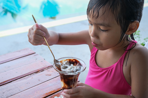 Wet little girl in a swimsuit eating ice cream by the outdoor pool during family summer vacation. Summer lifestyle concept.