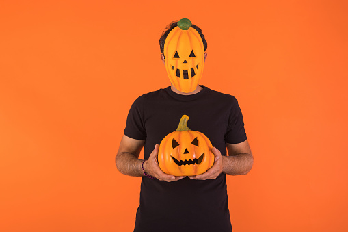 Person with pumpkin mask celebrating Halloween, holding a pumpkin, on orange background. Concept of celebration, All Souls' Day and All Saints' Day.