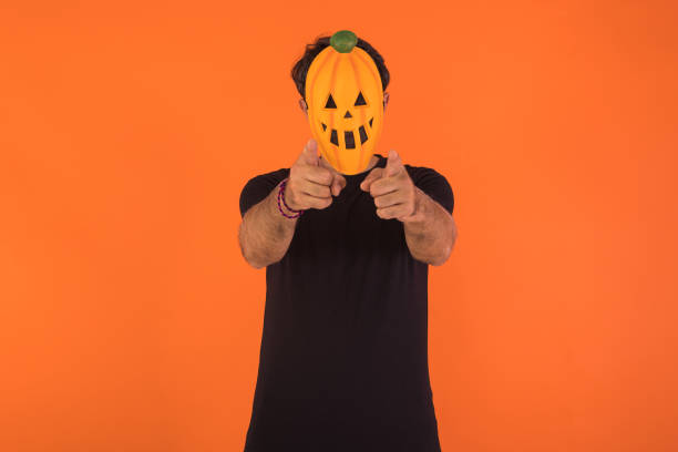 istock Person with pumpkin mask celebrating Halloween, pointing at camera, on orange background. Concept of celebration, All Souls' Day and All Saints' Day. 1429251069