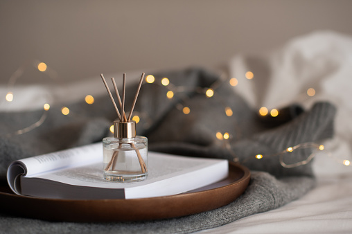 Liquid home fragrance stay on paper book with knit cloth sweater on wooden tray in bedroom in bed over glow christmas lights closeup. Cozy hygge atmosphere.