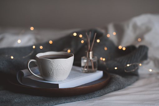 Cup of coffee on open paper book with liquid home perfume on wooden tray with christmas glow light in bed over knit cloth closeup. Cozy romantic atmosphere