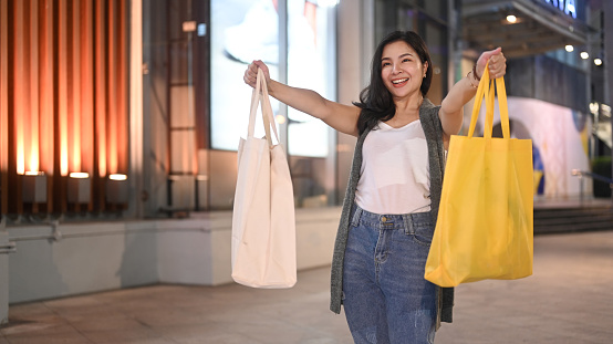 Happy asian woman carrying shopping bags standing at evening city streets. Black Friday, sale, shopping, lifestyle concept.