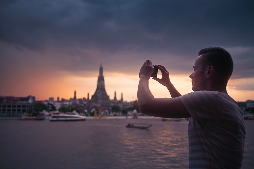 Man while taking pictures with mobile phone against Wat Arun temple. Tourist in Bangkok, Thailand.
