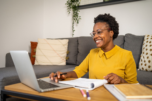 Smiling cute stylish African American female student with short hair, studying remotely from home, using a laptop, taking notes on notepad during online lesson, e-learning concept, smiling stock photo