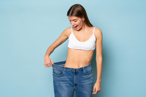 Woman astonished with results of her slimming and weight loss pulling large oversize jeans to check her success, isolated on blue background with wow face expression. Dieting, keep fit