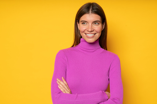 Portrait of confident woman office worker in pink turtleneck standing with crossed arms on yellow background looking aside thinking