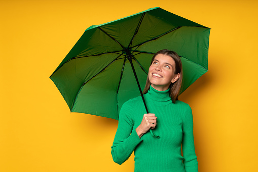 Studio portrait of dreamy romantic woman in green clothes hiding under umbrella on rainy day looking up on cloudy sky or rainbow on yellow background. Fall rain season. Weather forecast